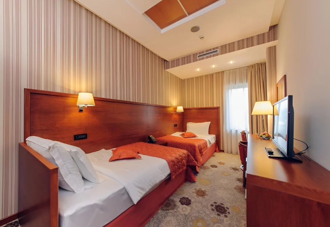 Cozy hotel room in Podgorica featuring two single beds with orange bedding, a large wooden cupboard with reading lamps and a flat-screen TV.