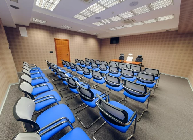 Spacious meeting room at Kings Park Hotel with rows of blue and black chairs, equipped with modern lighting and a projector screen, suitable for conferences and educational events.