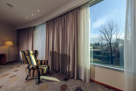 Hotel room in Podgorica with a large window offering a view of the outdoors, an elegant striped armchair, and soft ambient lighting, Kings Park Hotel.